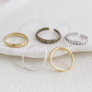 May include: Five open-ended rings in gold, silver, and bronze tones. The rings have a simple, minimalist design, with some featuring small, evenly spaced holes. The rings are displayed on a white surface, showcasing their varying metallic finishes.