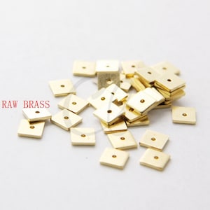 Puede incluir: Un montón de pequeñas piezas metálicas cuadradas de color latón con un agujero en el centro. RAW BRASS está escrito en letras rojas en el lado izquierdo de la imagen.