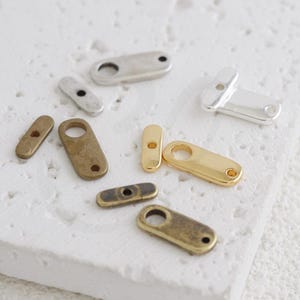 Op de afbeelding: Een verzameling kleine, langwerpige metalen sieradenverbinders in diverse afwerkingen, waaronder gepolijst goud, glanzend zilver, mat zilver en antiek messing. Elk stuk heeft een afgerond uiteinde met een rond gat en een kleiner gat aan de andere kant.