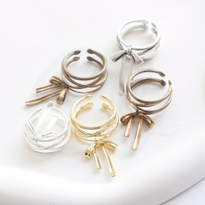 May include: Four silver, gold, and bronze rings with a bow detail. The rings are stacked on top of each other and are all different sizes. The rings are on a white background.