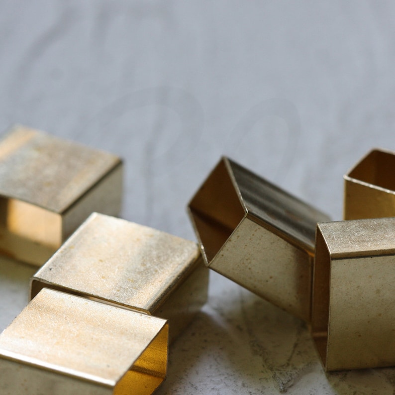 May include: A group of five small, gold-colored metal squares, some stacked on top of each other. The squares are hollow and have a slightly textured surface.