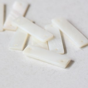 以下が含まれることがあります： 白い長方形の貝殻のピースがいくつかあり、それぞれに2つの穴が開いています。ピースは白い表面に積み重ねられています。