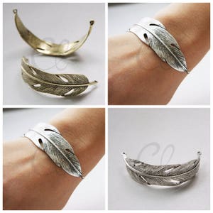May include: Two silver and two gold feather-shaped bracelets. The bracelets are open and have a delicate design. The bracelets are shown on a model's wrist and laid flat on a white surface.