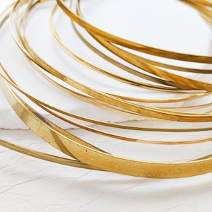 May include: Close-up of several gold-colored metal bangles, some with a flat, wide design, others thinner and round. The bangles are arranged in a swirling pattern, creating a layered effect. The background is white.