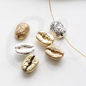 Può includere: Una collezione di sei ciondoli a forma di chicco di caffè dorati e argentati. I ciondoli sono testurizzati e hanno una forma realistica di chicco di caffè.