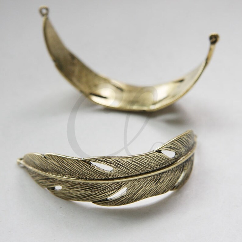 May include: Two gold-toned metal feather-shaped charms with cut-out details. The charms are curved and have a delicate, intricate design.