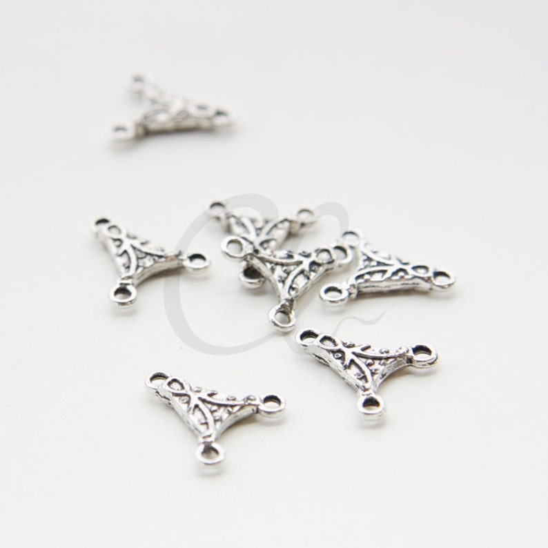 May include: Close-up of several silver-colored, ornate metal connectors. Each connector has a triangular shape with three loops and decorative filigree patterns. The connectors are scattered on a white surface.