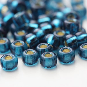 May include: A close-up of a pile of teal blue glass beads with a metallic sheen. The beads are round and have a small hole in the center.