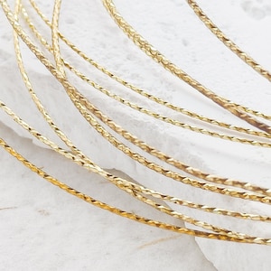 May include: Close-up of several strands of gold-coloured wire. The wire has a textured, faceted surface, catching the light and creating a shimmering effect. The wires are arranged in a slightly curved pattern against a white background.