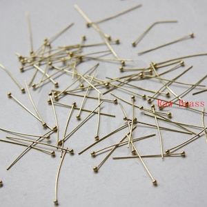 May include: A large quantity of gold-colored head pins with round heads scattered on a white surface. The text "Raw Brass" is visible in the lower right corner of the image.