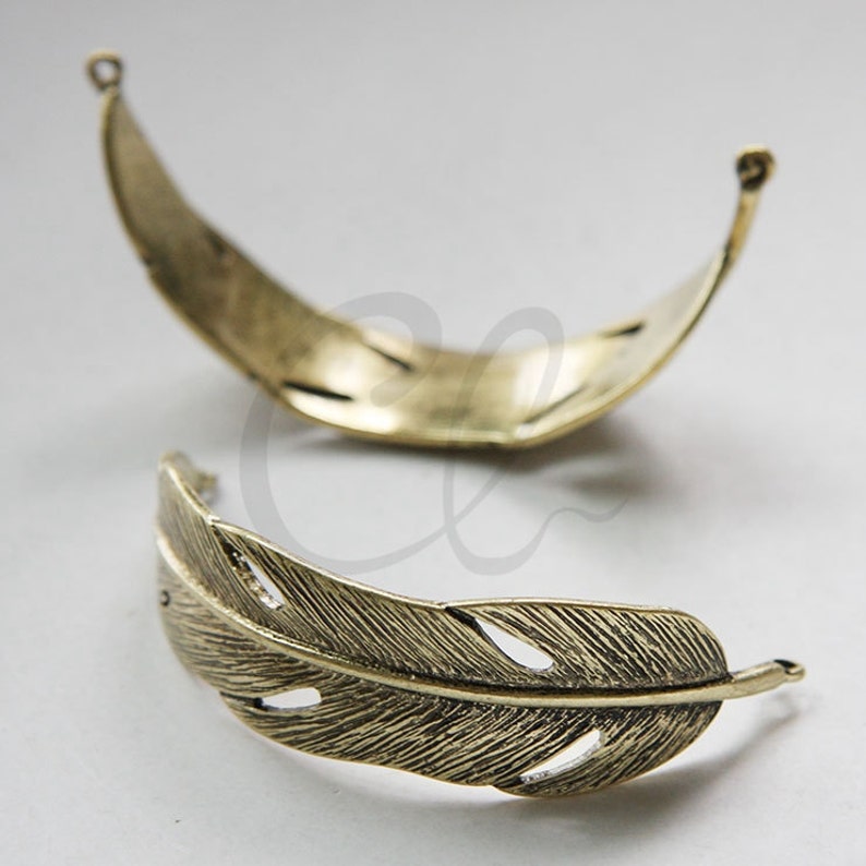 May include: Two gold-toned metal feather-shaped charms. The charms are open on one side and have a detailed feather design.