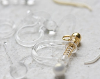 May include: A close-up of a clear plastic earring back with a gold spring and a pearl-like bead. The earring back is designed to be used with a variety of earrings.