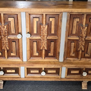 May include: A wooden sideboard featuring three upper cabinet doors and three lower drawers. The doors and drawer fronts display a geometric pattern with carved central panels. The piece includes light grey vertical accents and round white knobs.