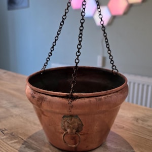 May include: A copper-toned hanging planter with a lion's head ring detail. It is suspended by a dark metal chain. The planter is on a wooden table.