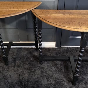 May include: Two wooden half-moon tables. The tabletops are light brown with a natural wood grain. The legs are black, with a twisted, rope-like design. The tables are on a dark grey carpet.