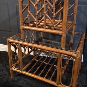 May include: A two-tiered, vintage bamboo side table with a glass top. The table has a natural wood finish and features intricate woven detailing on the sides and top shelf. The lower level has a slatted shelf for storage. The table is approximately 61 cm tall.