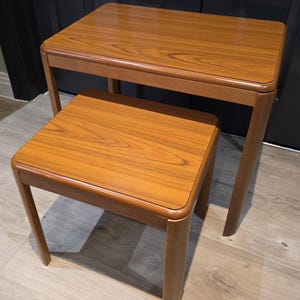 May include: A set of two rectangular nesting tables with a warm brown wood grain finish. Each table features rounded corners on the tabletop and four sturdy legs. The smaller table is positioned in front of the larger table, both showing a smooth surface.