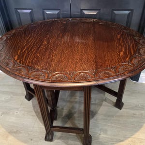 May include: A round, dark brown wooden table with a prominent grain pattern. The tabletop features a carved border with repeating fan-like motifs. The table is supported by four vertical, fluted legs connected by a lower rectangular frame.