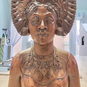 May include: A detailed wooden sculpture of a woman with closed eyes, adorned with an elaborate headdress and ornate jewellery. The statue has a warm, brown tone and is likely a decorative piece or a work of art.
