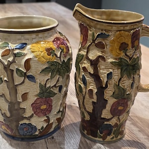 May include: A decorative vase and jug set. The beige ceramic pieces feature a raised floral and tree design with yellow, red, blue, and green accents. The jug has a handle and spout.