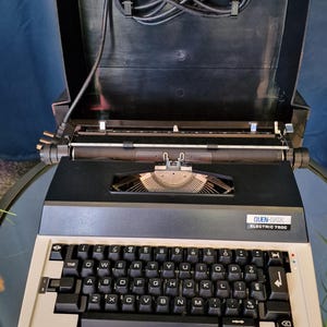 May include: A vintage Quen-Vox Electric 750C typewriter in black and beige. It features a full keyboard, a carriage, and a black carrying case. The text "QUEN-VOX ELECTRIC 750C" is displayed on the front of the machine.