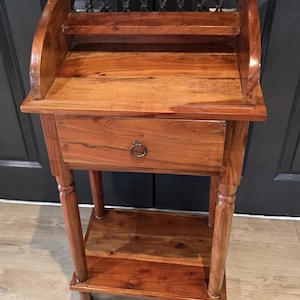 May include: A brown wooden side table featuring a curved top gallery with a decorative metal lattice insert, a single drawer with a circular metal pull, and a lower shelf supported by four turned legs with a warm, polished wood grain finish.