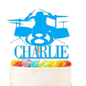 May include: A blue cake topper featuring a drummer silhouette, the number 8, and the name "Charlie." The cake is white with colorful sprinkles and decorative frosting.