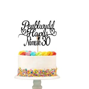 Puede incluir: Topper para pastel de acrílico negro con el texto "Penblwydd Hapus Nimesh 30" y dos corazones. El topper está sobre un pastel blanco con chispas de colores.