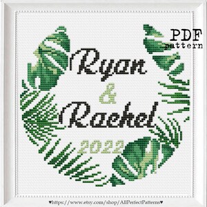 May include: Cross stitch pattern featuring a green leafy wreath with the names "Ryan & Rachel" and the year "2022" in the center. The pattern is labeled "PDF pattern".