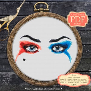 Peut inclure: Un motif de point de croix représentant deux yeux avec du maquillage rouge et bleu, encadré dans un cercle à broder en bois. Le motif comprend un petit cœur noir et le texte "Cross Stitch PDF Pattern". La taille du motif est de 130w X 55h points.