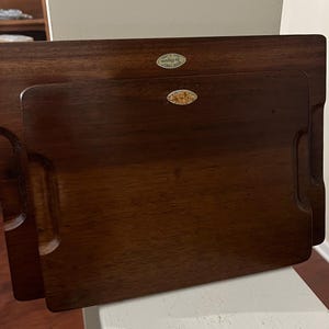 May include: Two rectangular wooden serving trays with carved handles. The trays are a rich, dark brown color and have a smooth finish. Each tray has a small, oval sticker. The trays are nested together, with the smaller tray inside the larger one.