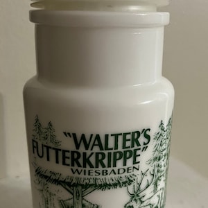 Puede incluir: Jarrón de cerámica blanca con un diseño verde y blanco. El jarrón tiene una tapa y presenta el texto "Walter's Futterkrippe" y "Wiesbaden". El diseño incluye una pequeña estructura de madera y ciervos.