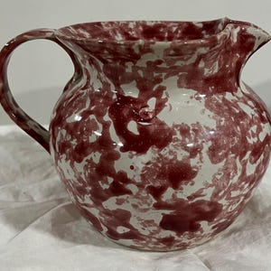 May include: A ceramic pitcher with a rounded body, a handle, and a pouring spout. The pitcher is white with a red splatter pattern. The handle is curved and the spout is slightly pointed. The pitcher is likely used for serving beverages.