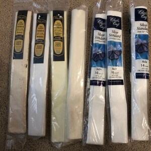 May include: Eight packages of Aida fabric for cross stitch. The packages are labeled with the count, color, and size of the fabric. The fabric is white, ivory, and antique white. The packages are labeled with the brand name Charles Craft.