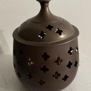 May include: A dark brown metal incense burner with a lid and a decorative design. The burner has a round body with a series of small, open circles around the sides. The lid has a small, round knob on top.