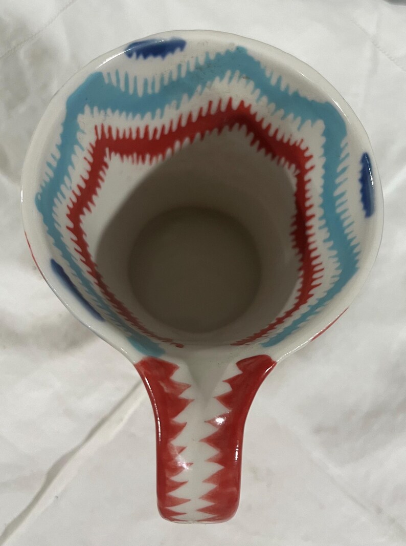 May include: White ceramic pitcher with a red and blue zigzag pattern around the rim and on the handle. The interior is plain. The pitcher is designed for serving beverages.