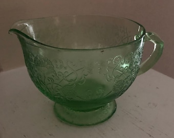 May include: A green glass creamer with a floral design. The creamer has a handle and a pedestal base.