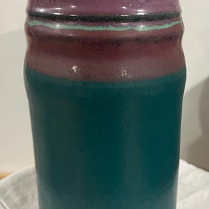 May include: A cylindrical ceramic vase featuring a smooth, matte teal base that transitions into a wide band of deep plum purple. The upper rim is accented with a thin, horizontal stripe of light mint green and a dark charcoal line, finished with a blue-toned edge.