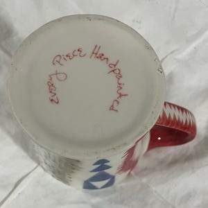 May include: Hand-painted ceramic mug with a red handle and geometric patterns in red, blue, and gray. The bottom of the mug has the text "Every Piece Handpainted" in red script.