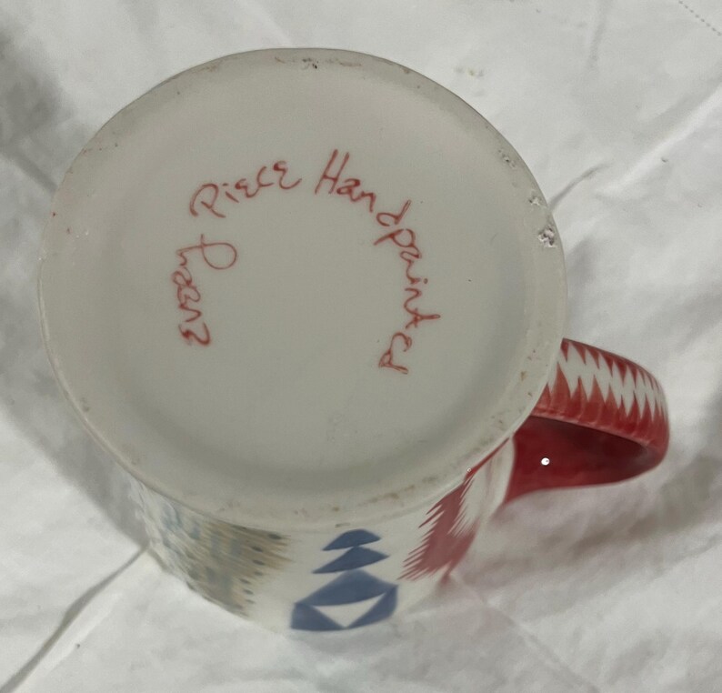 May include: Hand-painted ceramic mug with a red handle and geometric patterns in red, blue, and gray. The bottom of the mug has the text "Every Piece Handpainted" in red script.