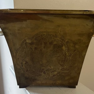 May include: A large, brass planter with an ornate, embossed design featuring a scene of birds and trees. The planter has a square base and flared sides with two small handles.