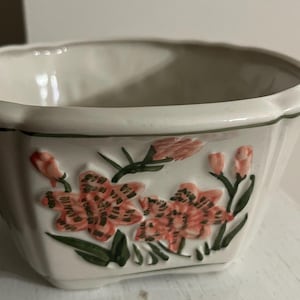 May include: A white ceramic bowl with a scalloped rim and a floral design. The flowers are pink and orange with green leaves. The bowl has a square shape with slightly angled sides.