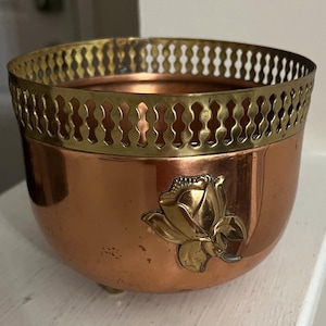 May include: A copper-colored bowl with a brass rim and a brass rose applique. The bowl has a rounded shape and sits on three small brass feet. The rim features a decorative pierced design.