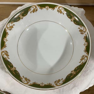 May include: A round white ceramic bowl featuring a wide rim decorated with a green band and gold filigree scrollwork patterns. The center of the bowl is plain white, and the outer edge is finished with a thin gold trim along the circumference.