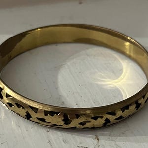 May include: A gold-colored bangle bracelet with a floral cutout design. The bracelet is a solid band with a repeating pattern of stylized flowers and leaves around the lower edge. The metal has a slightly aged appearance.