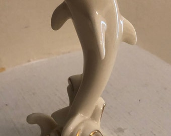 May include: A white porcelain figurine of a dolphin leaping out of the water. The dolphin is painted with gold accents and has a detailed, realistic design.