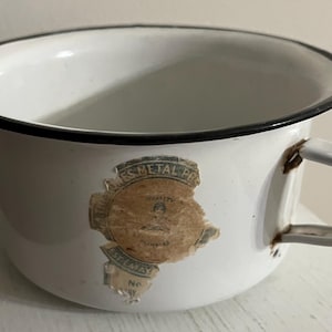 May include: White enamelware cup with a black rim and handle. The cup has a faded label with text. The handle is black and white. A vintage kitchenware item.