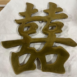 May include: A polished brass wall hanging featuring the Chinese double happiness character. The metal object has a smooth, reflective surface with a warm gold tone and includes a small circular loop at the top center for mounting purposes.