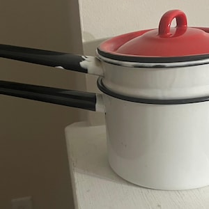 May include: White enamel double boiler with a red lid and black handles. The pot has a classic design, with a rounded body and a lid with a loop handle. The handles are long and angled. The pot is likely used for cooking.