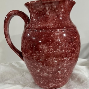 May include: A maroon ceramic pitcher with a speckled white pattern. The pitcher has a curved handle and a flared spout. The surface is glossy. The pitcher is likely used for serving beverages.